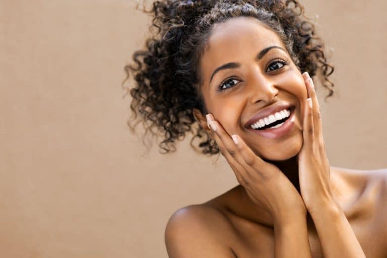 Image starts just below collarbone of a happy woman with dark hair and eyes with each hand placed on the corresponding cheek. Her mouth is open in a smile and she appears to be delighted. 